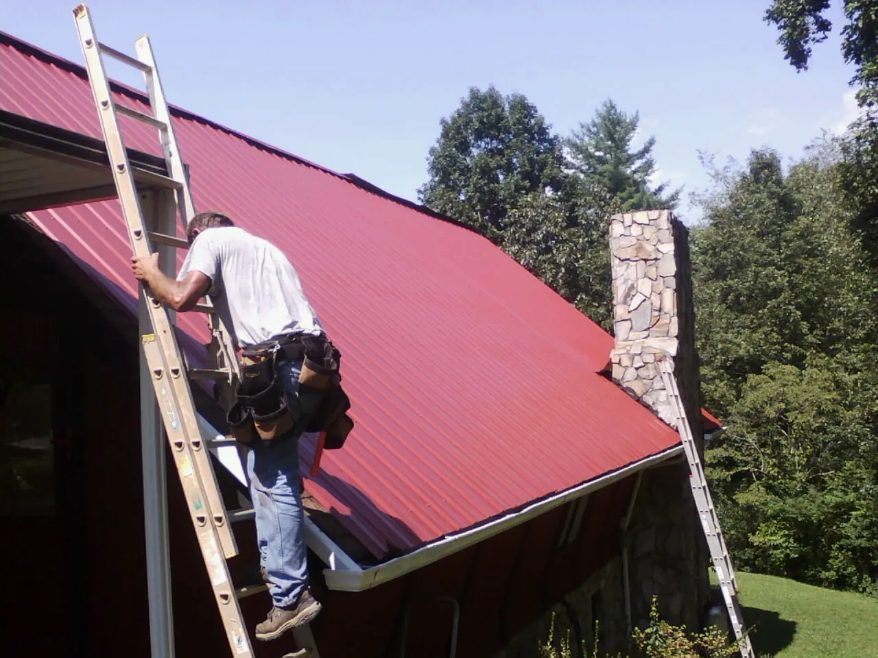 Roofer on a ladder beside a red metal roof and stone chimney — Soffit & Fascia Repair by Roofing Brothers in New Caney